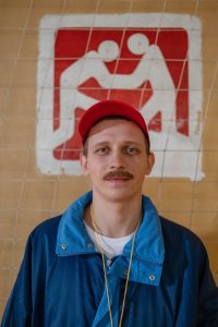 Image d'un entraîneur sportif masculin avec une moustache, portant une veste bleue et une casquette rouge, debout à l'intérieur.