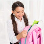 Une jeune fille souriante avec des couettes ouvre son sac à dos Barbie rose pour y glisser des cahiers et fournitures scolaires.